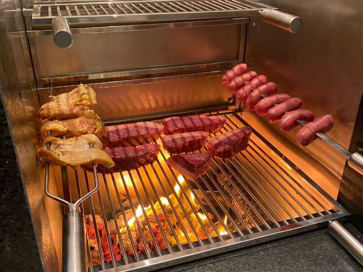 Espetos de inox com frango e linguiça assando sobre a brasa, ao lado da picanha na grelha