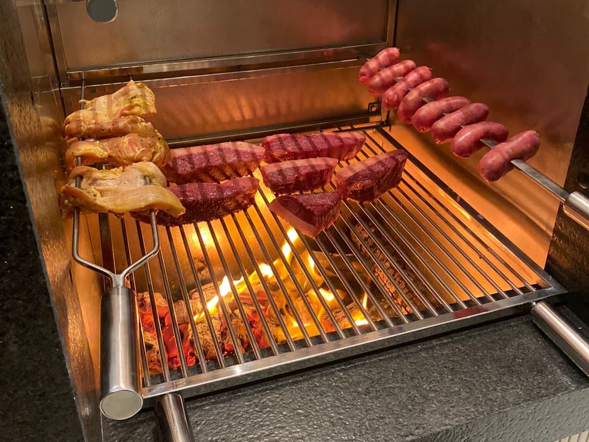 A churrasqueira em uso, com carne na grelha e espetos de linguiça e frango presos no suporte aéreo