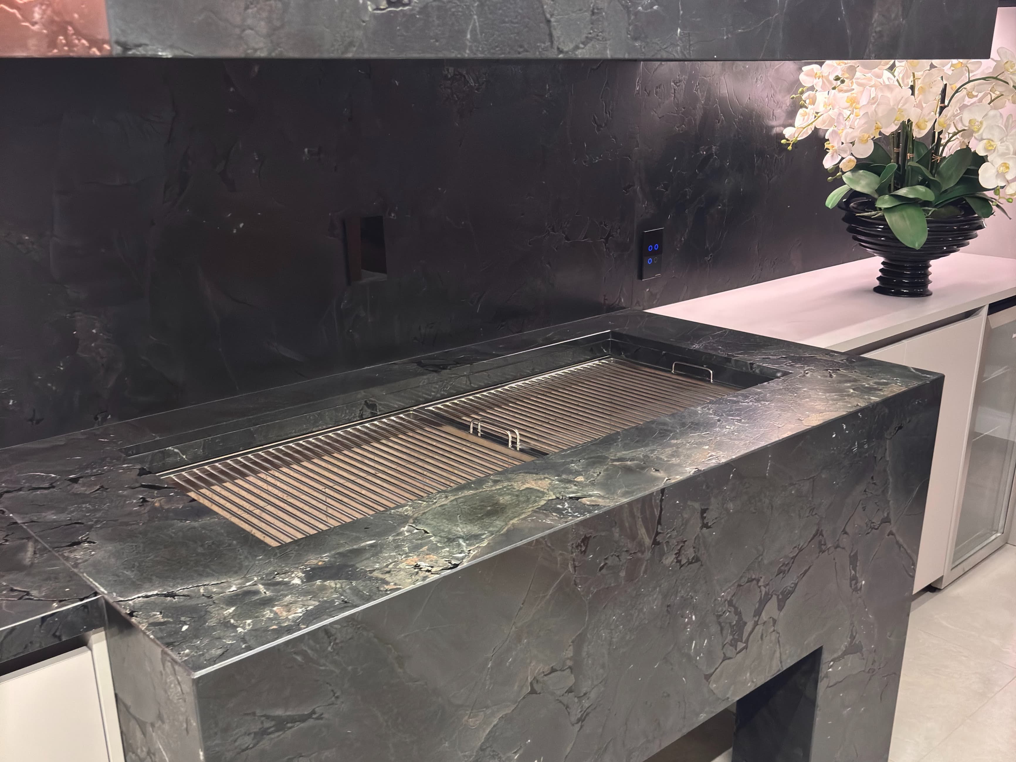 Churrasqueira de inox embutida em bancada de pedra escura, em área gourmet com parede no mesmo revestimento